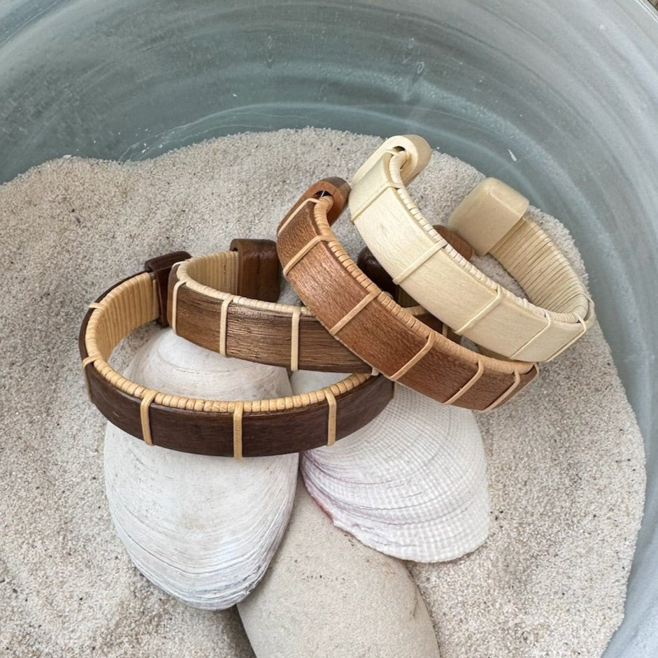 Four Boardwalk cuff bracelets in cherry, holly, rosewood, and walnut displayed on seashells in sand, showcasing the open-weave cane design and available wood variations.