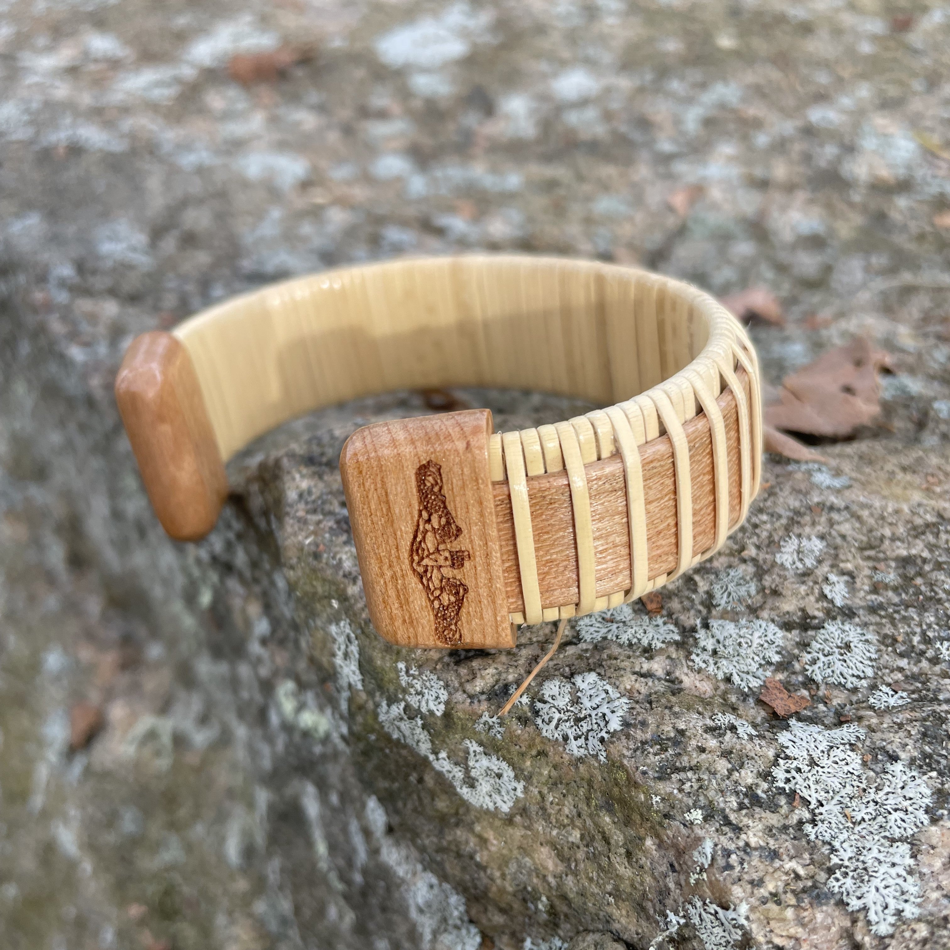 Handcrafted cuff bracelet with light weaver detail and a cherry wood end cap etched with a submarine design, resting on a rock.