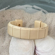Holly Beam cuff bracelet resting on seashells and a smooth stone in sand, showing the bold open-weave cane design and natural pale wood tone.