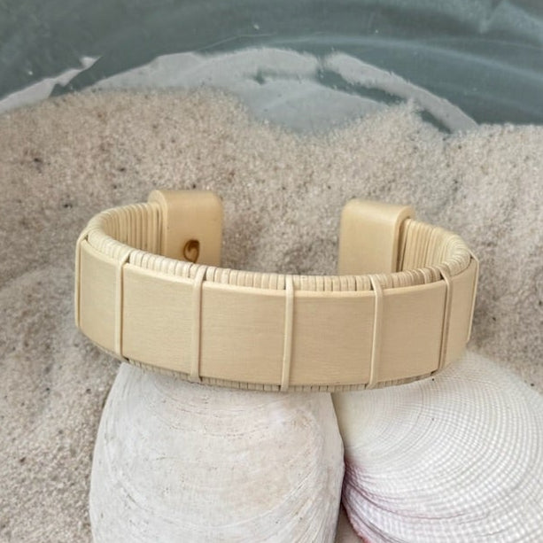 Holly Beam cuff bracelet resting on seashells and a smooth stone in sand, showing the bold open-weave cane design and natural pale wood tone.