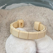 Holly Boardwalk cuff bracelet resting on seashells and a smooth stone in sand, showing the open-weave cane design and pale natural wood tone.
