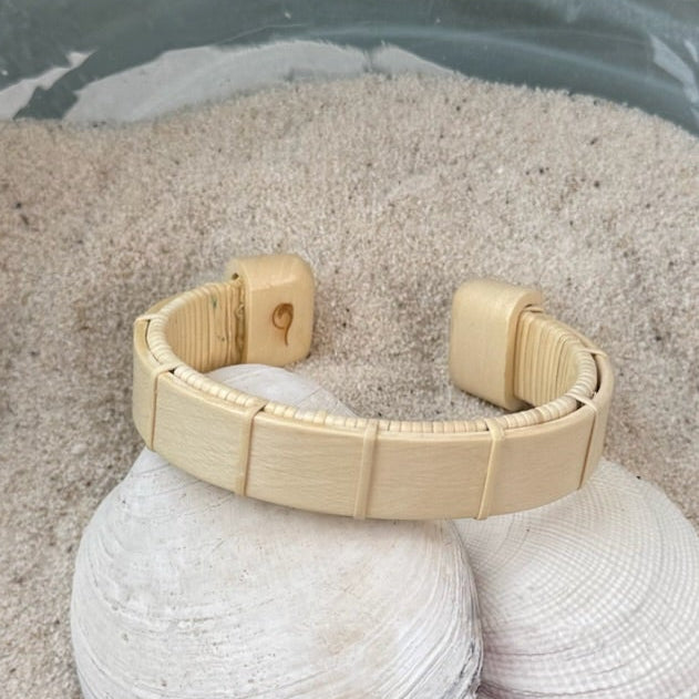 Holly Boardwalk cuff bracelet resting on seashells and a smooth stone in sand, showing the open-weave cane design and pale natural wood tone.