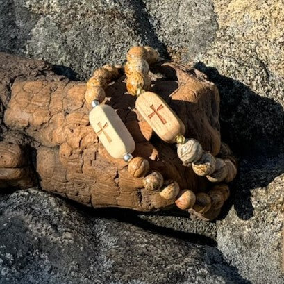 Close-up of two handcrafted Slivers cross bracelets with etched holly and cherry wood beads paired with picture jasper stone beads, resting on driftwood.