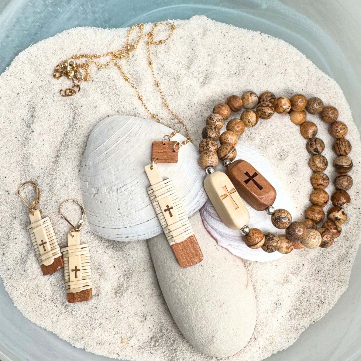 Handcrafted Slivers cross collection featuring earrings, a necklace, and beaded bracelets with etched holly and cherry wood accents, displayed in sand with a shell.