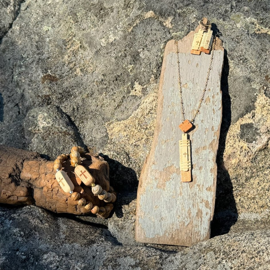 Handcrafted Slivers cross collection styled on rocks, with beaded bracelets, a necklace, and earrings displayed on a driftwood stand.