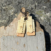 Close-up of handcrafted Slivers cross earrings with woven holly wood and cherry wood base, hanging against a rustic driftwood and rock backdrop.