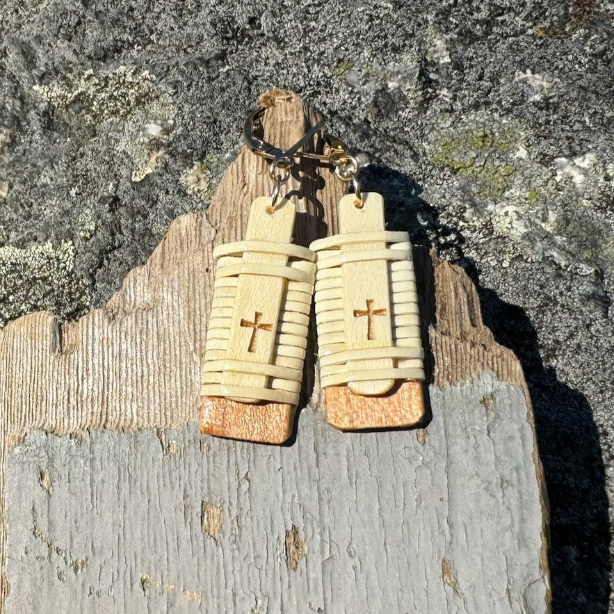 Close-up of handcrafted Slivers cross earrings with woven holly wood and cherry wood base, hanging against a rustic driftwood and rock backdrop.