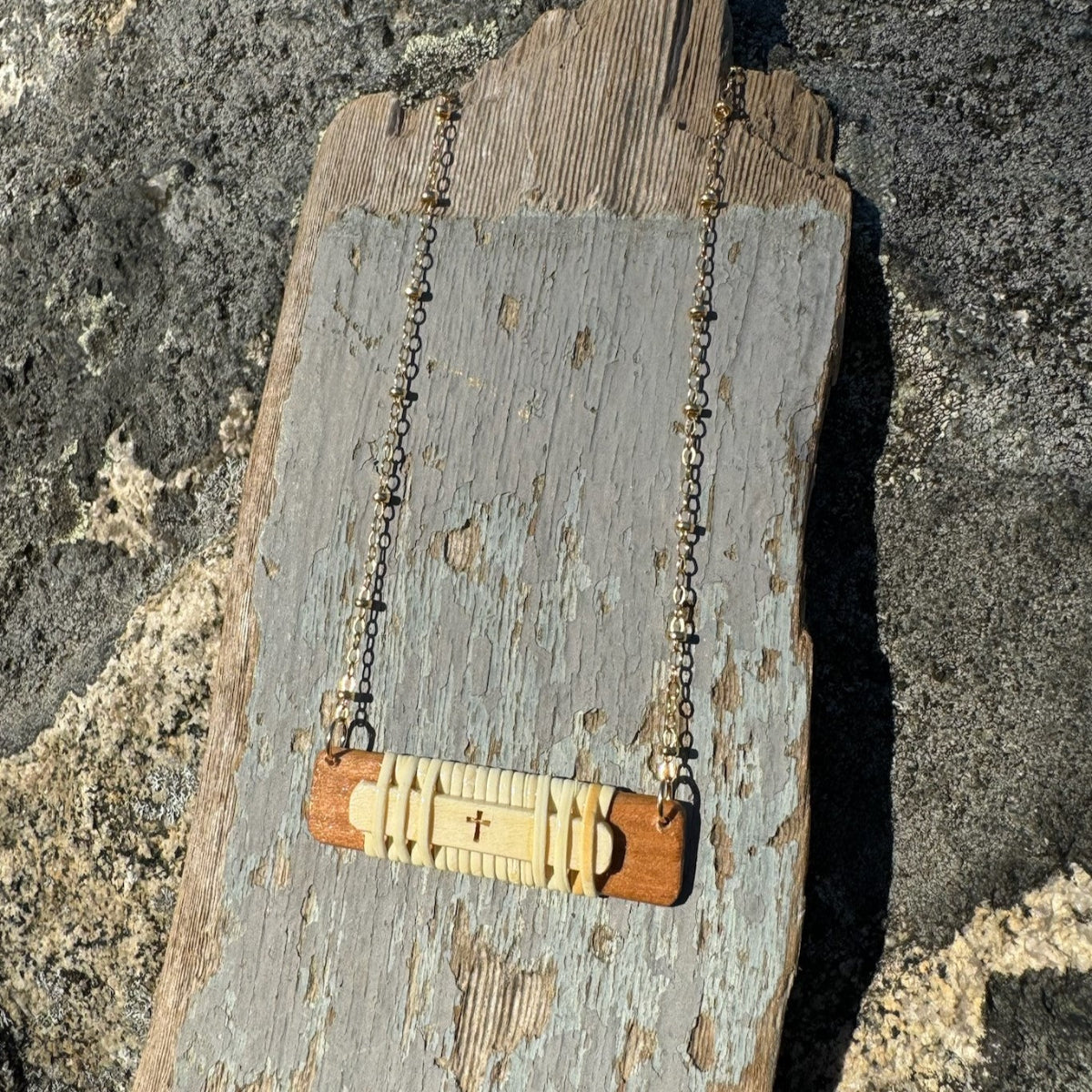 Handcrafted horizontal Slivers cross necklace with woven holly wood and cherry wood base, draped on a chain over a piece of driftwood.