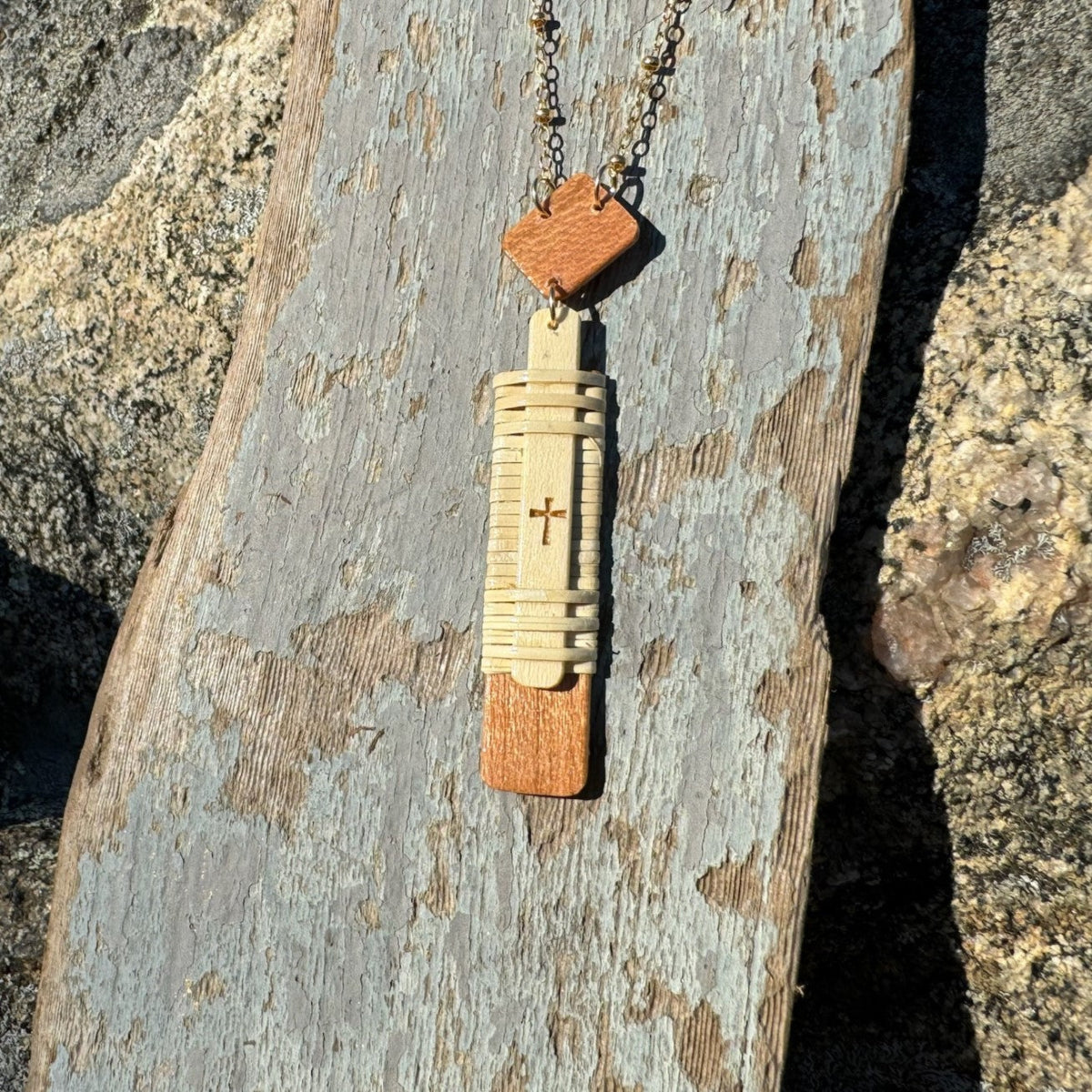 Handcrafted vertical Slivers cross necklace with woven holly wood and cherry wood base, draped on a piece of driftwood.