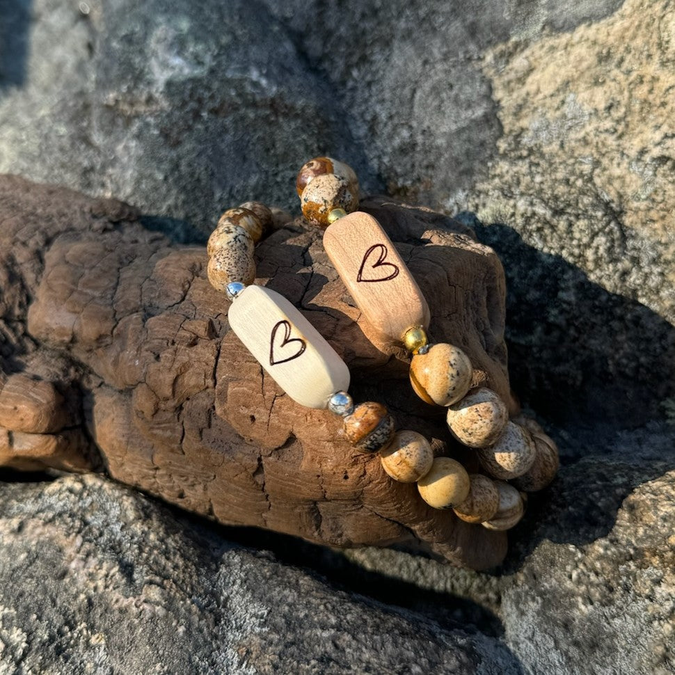Close-up of two handcrafted Slivers heart bracelets with etched holly and cherry wood beads paired with picture jasper stone beads, resting on driftwood.