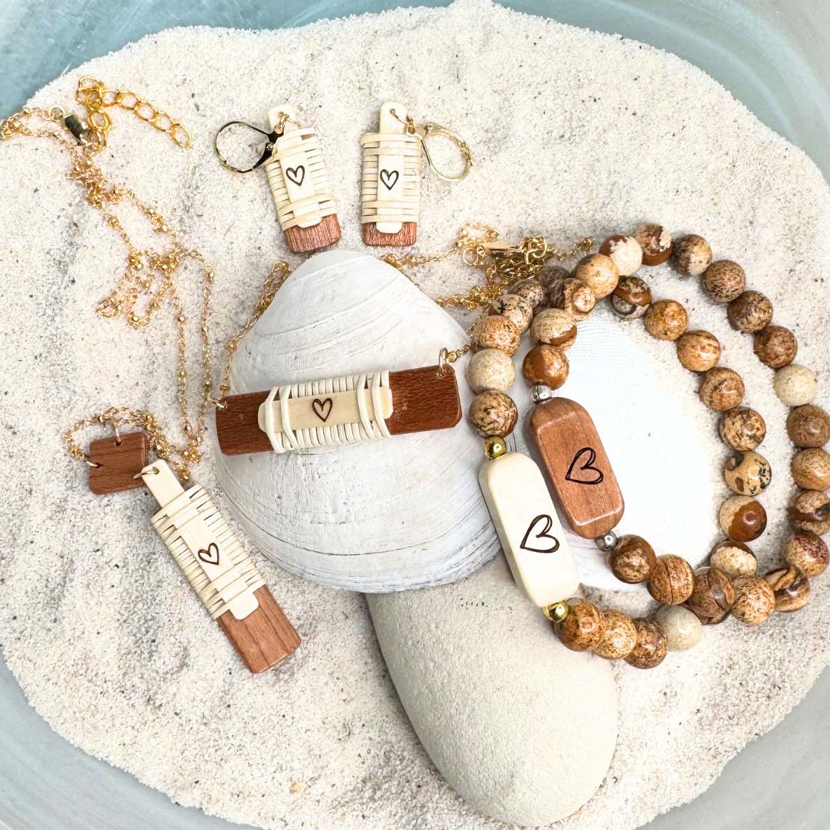 Handcrafted Slivers heart collection featuring earrings, a necklace, and beaded bracelets with etched holly and cherry wood accents, displayed in sand with a shell and stone.