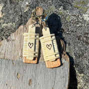 Close-up of handcrafted Slivers heart earrings with woven holly wood and cherry wood base, hanging against a rustic driftwood and rock backdrop.