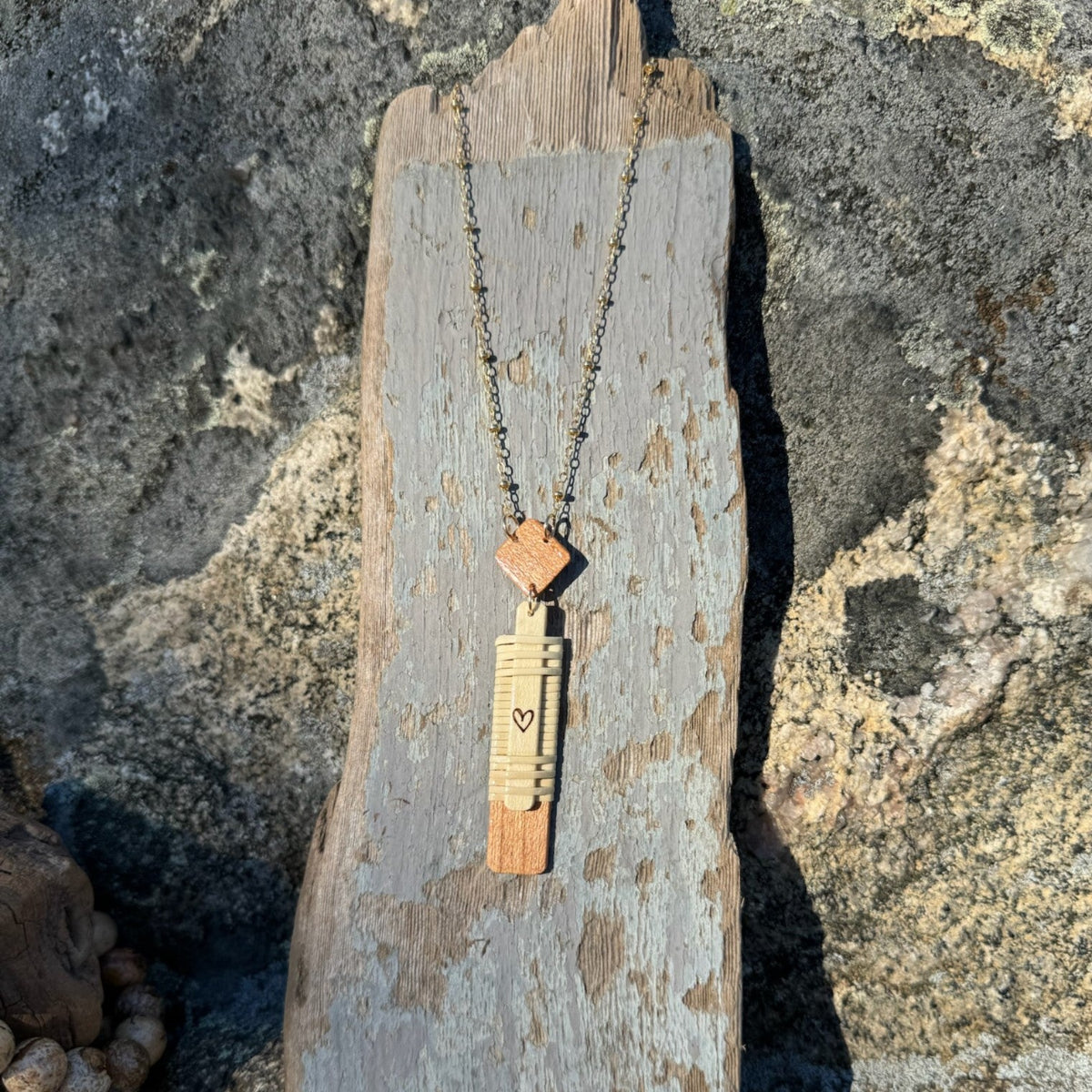 Handcrafted vertical Slivers heart necklace with woven holly wood and cherry wood base, draped on a piece of driftwood.