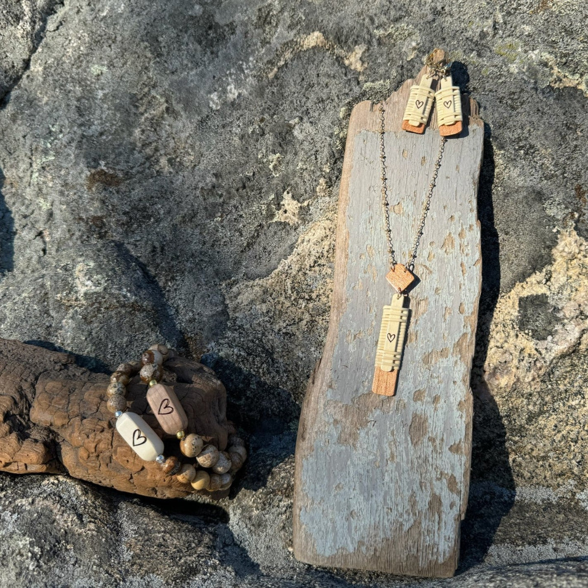 Handcrafted Slivers heart collection styled on rocks, with a vertical necklace, beaded bracelets, and earrings displayed on a driftwood stand.