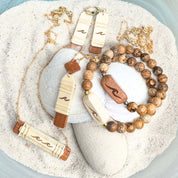 Handcrafted Slivers wave collection featuring earrings, a necklace, and beaded bracelets with etched holly and cherry wood accents, displayed in sand with a shell.