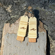 Close-up of handcrafted Slivers wave earrings with woven holly wood and cherry wood base, hanging against a rustic driftwood and rock backdrop.
