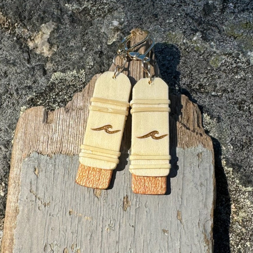 Close-up of handcrafted Slivers wave earrings with woven holly wood and cherry wood base, hanging against a rustic driftwood and rock backdrop.