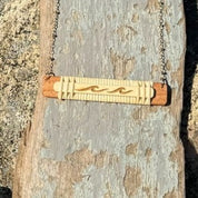 Handcrafted horizontal Slivers wave necklace with two etched waves on a woven holly wood and cherry wood base, draped on a piece of driftwood.