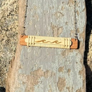 Handcrafted horizontal Slivers wave necklace with two etched waves on a woven holly wood and cherry wood base, draped on a piece of driftwood.