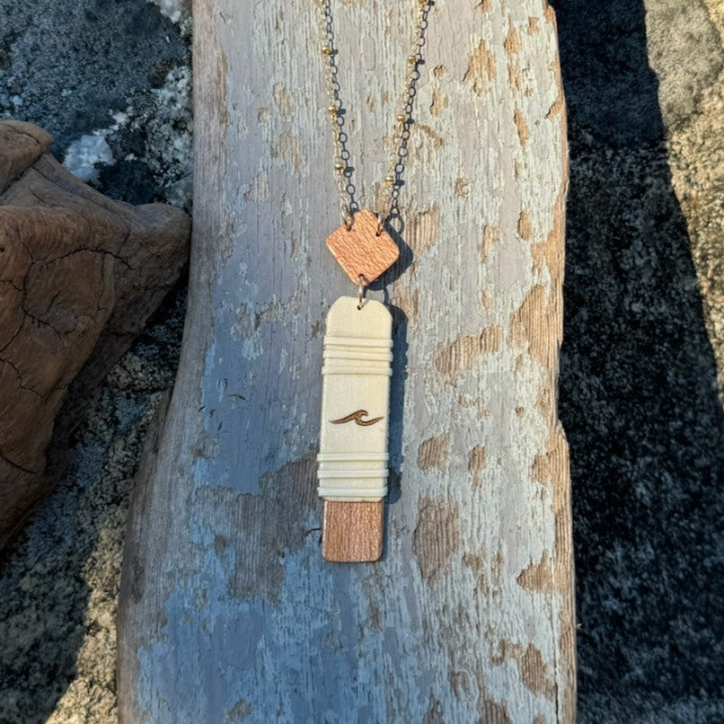 Handcrafted vertical Slivers wave necklace with a single etched wave on a woven holly wood and cherry wood base, draped on a piece of driftwood.