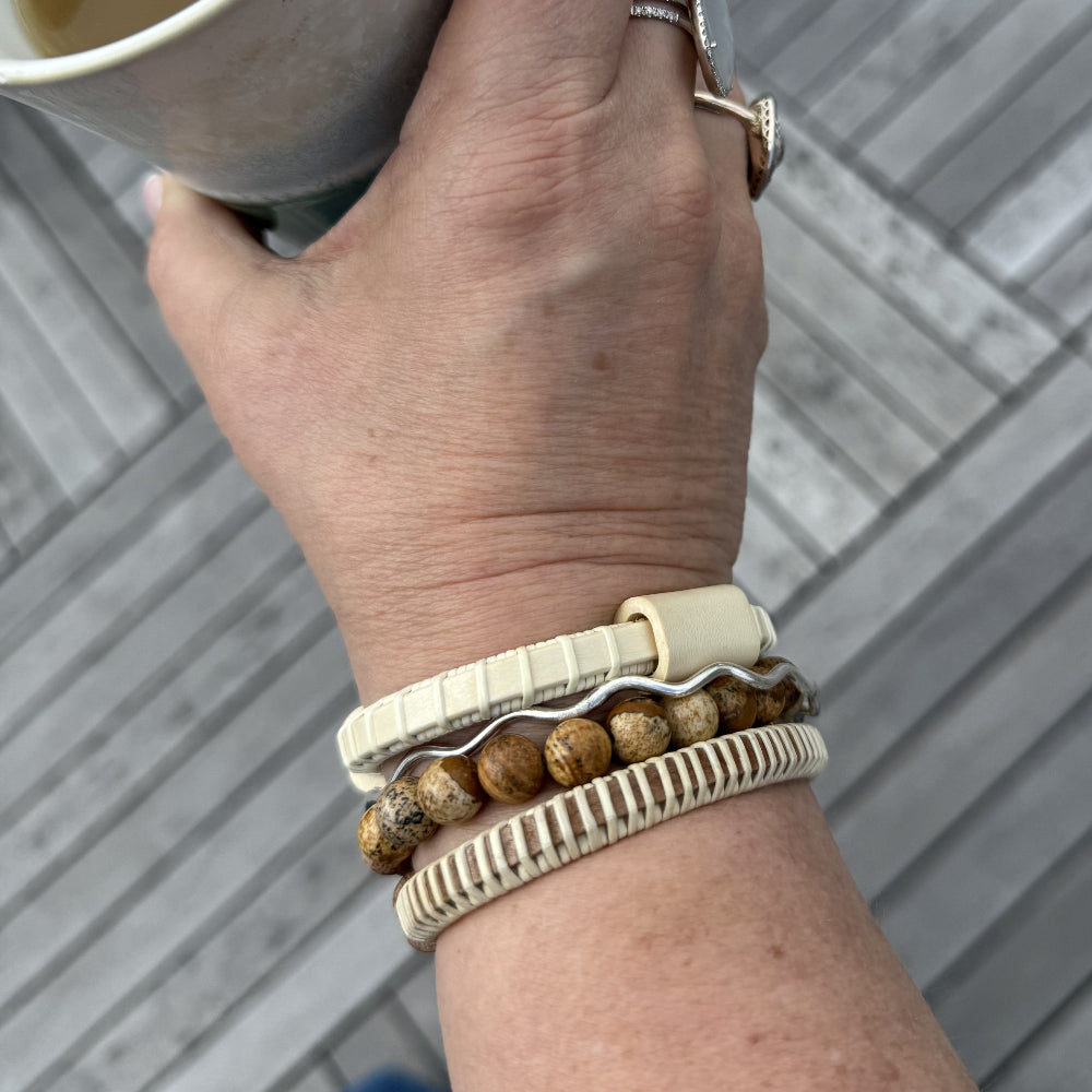 Holly Cove cuff bracelet with holly slider worn stacked on a wrist with other bracelets, hand holding a cup on a wooden deck background.