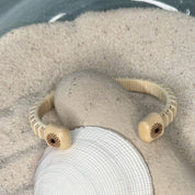 Handcrafted holly Little Daisies Etched Cuff Bracelet with daisy-engraved holly end caps resting on a smooth stone and seashell in sand, made by military spouse artisans at Stonington Designs.