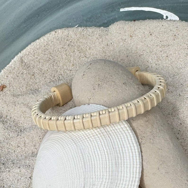 Side angle of the handcrafted holly Little Daisies Etched Cuff Bracelet resting on a smooth stone and seashell in sand, showing the fine holly weave construction detail.