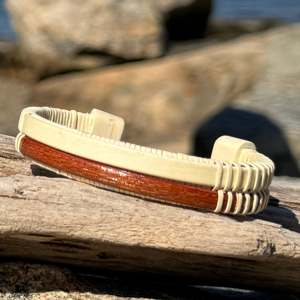 Handcrafted open weave cuff bracelet pairing light holly wood with a mahogany center panel, resting on driftwood with rocks and water in the background.