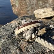 Handcrafted open weave cuff bracelet pairing light holly wood with a rosewood center panel, hanging on driftwood with water in the background.