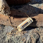 Handcrafted Patriot Necklace with a horizontal bar pendant in holly, white oak, and navy bamboo on a gold-filled chain, displayed on driftwood and coastal rocks, part of the America250 Collection by Stonington Designs.