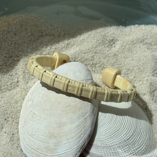 Handcrafted holly wood Clubhouse cuff bracelet with light weaver detailing, resting on a seashell in the sand — handmade by Military Spouse artisans in Stonington, Connecticut using American-made materials.
