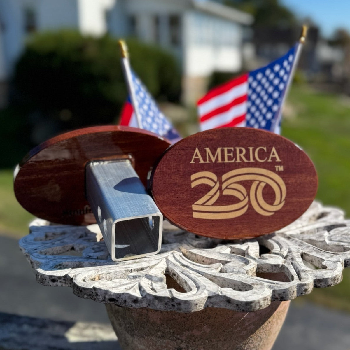 Front and back view of a mahogany and maple America 250 hitch cover with metal post, displayed outdoors with two American flags in the background