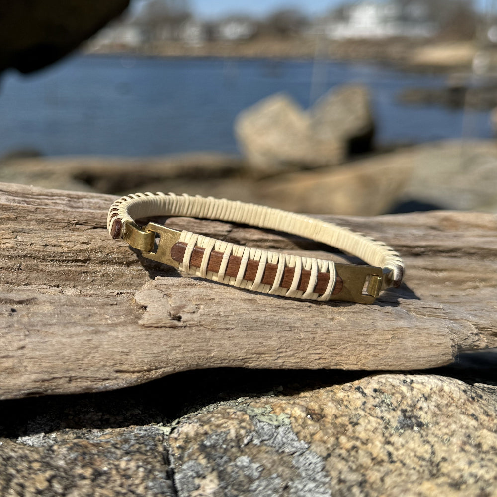 Handcrafted mahogany Nauti Clasp Bracelet with brass clasp resting on driftwood and coastal rocks with water visible in the background.