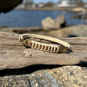 Handcrafted mahogany Nauti Clasp Bracelet with brass clasp resting on driftwood and coastal rocks with water visible in the background.
