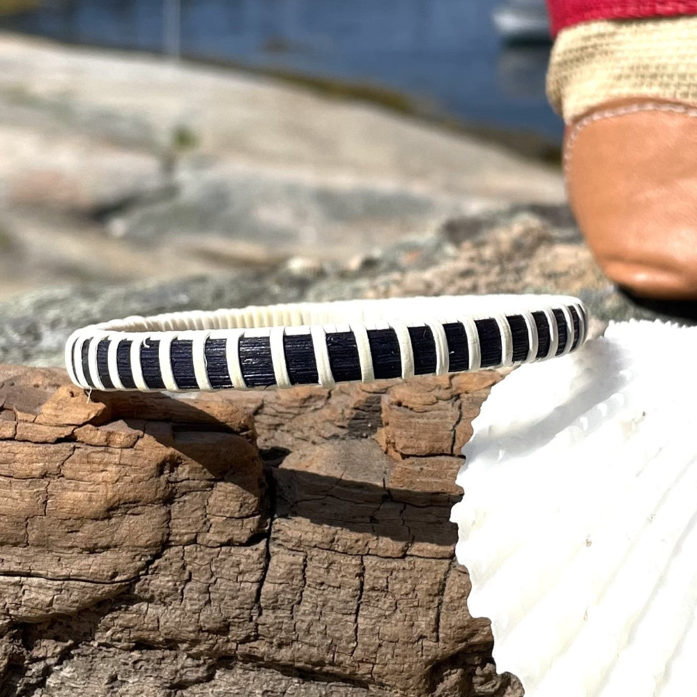Single navy and natural cane bangle bracelet resting on driftwood beside a seashell, with a harbor visible in the background