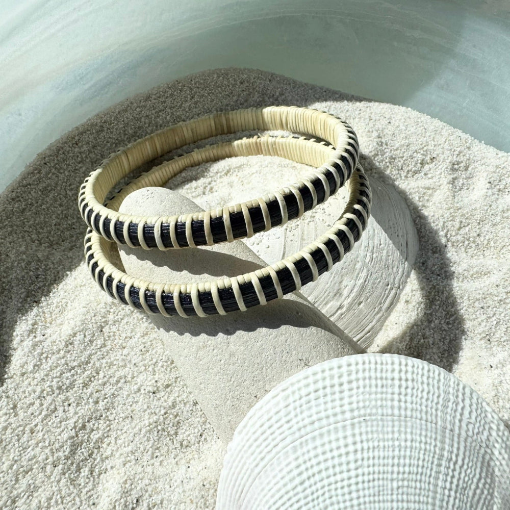 Two navy and natural cane Americana bangle bracelets stacked on a smooth stone in sand, with a seashell nearby