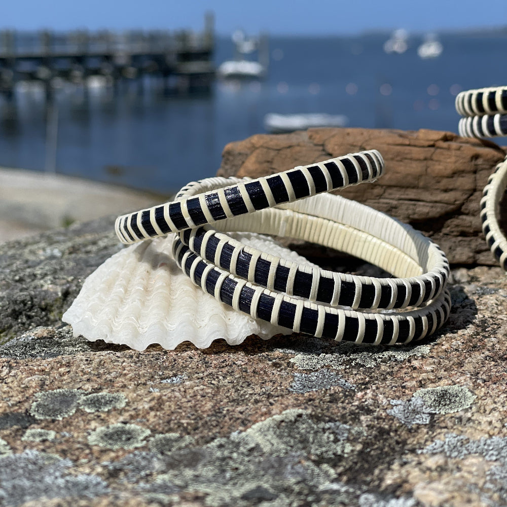 Two navy and natural cane bangle bracelets stacked on a seashell atop granite rocks, with a marina and blue sky in the background