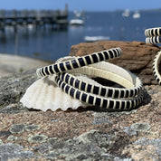 Two navy and natural cane bangle bracelets stacked on a seashell atop granite rocks, with a marina and blue sky in the background