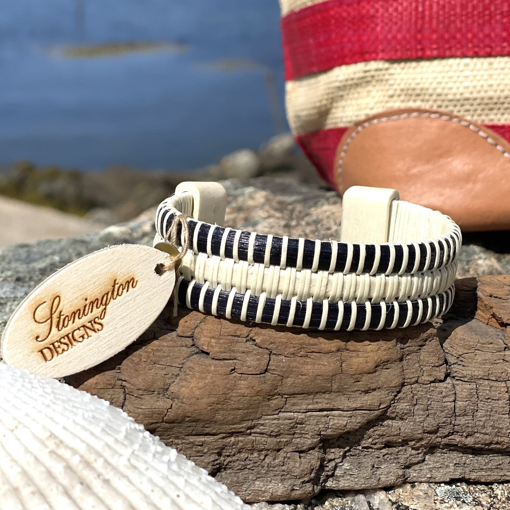 Handcrafted Lineage Triple Cuff Bracelet in navy cane and holly resting on driftwood with a Stonington Designs hang tag, seashell, and coastal water in the background, made by military spouse artisans.