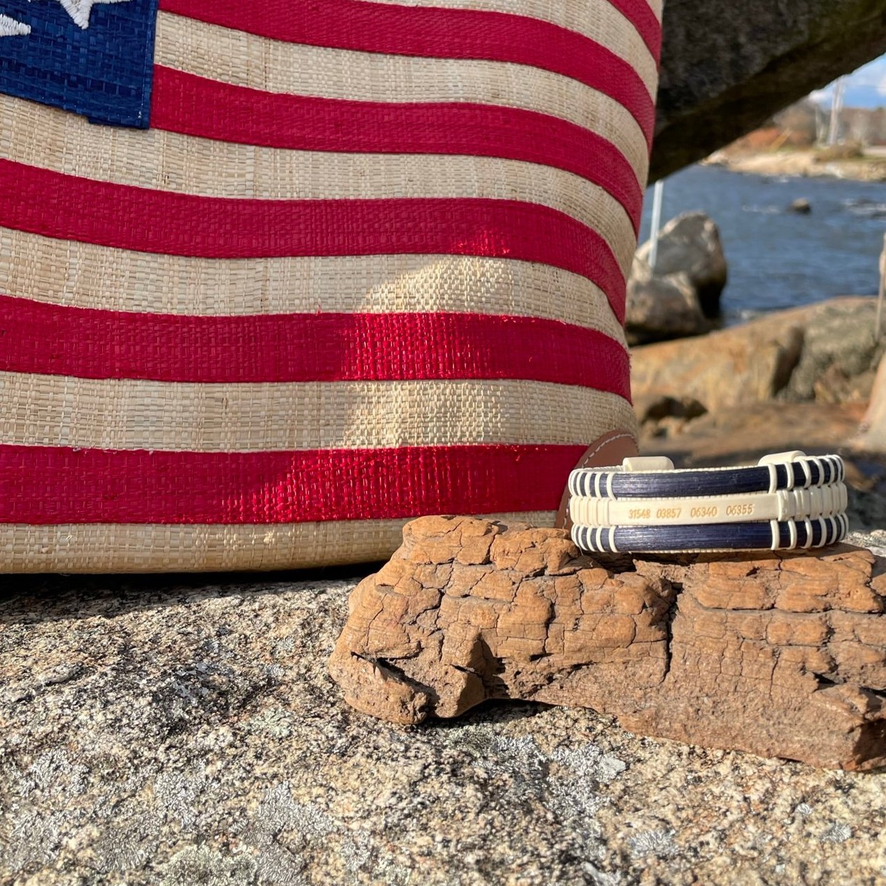 Handcrafted navy cane and holly PCS Seasons Etched Triple Cuff Bracelet with latitude and longitude coordinates etched on the holly stave, resting on driftwood in front of an American flag tote bag on coastal rocks, a meaningful personalized gift for military families.