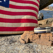 Handcrafted navy cane and holly PCS Seasons Etched Triple Cuff Bracelet with latitude and longitude coordinates etched on the holly stave, resting on driftwood in front of an American flag tote bag on coastal rocks, a meaningful personalized gift for military families.