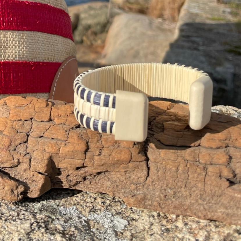 Handcrafted navy cane and holly PCS Seasons Etched Triple Cuff Bracelet resting on driftwood against coastal rocks with a red-and-white tote bag in the background, showing the wide triple-stave construction.