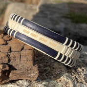 Close-up of the handcrafted navy cane and holly PCS Seasons Etched Triple Cuff Bracelet resting on a rock, showing latitude and longitude coordinates etched on the middle holly stave as a personalized military move keepsake.
