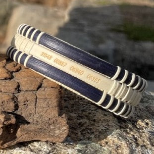 Close-up of the handcrafted navy cane and holly PCS Seasons Etched Triple Cuff Bracelet resting on a rock, showing latitude and longitude coordinates etched on the middle holly stave as a personalized military move keepsake.