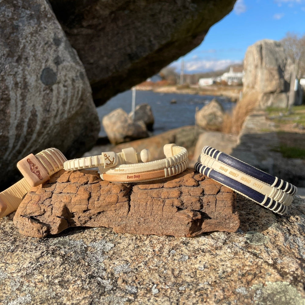 Handcrafted PCS Seasons Etched Triple Cuff Bracelet displayed on driftwood alongside the Our Love Runs Deep Double Cuff and a cherry cuff bracelet, with coastal rocks and water in the background, showing Stonington Designs military-themed bracelet collection.