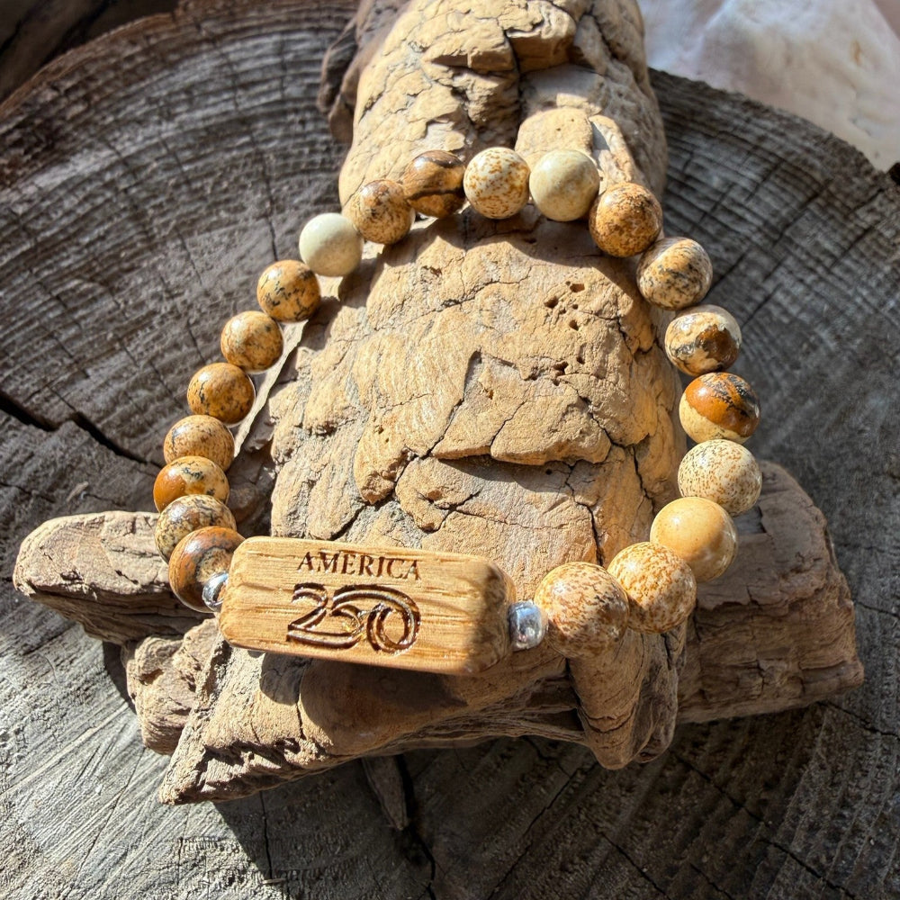 Handcrafted Old Glory Bead Bracelet with Picasso jasper stone beads and a white oak bead etched with the America250 logo, draped over driftwood on a wood stump, part of the licensed America250 Collection by Stonington Designs.