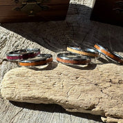 Handcrafted Whisper 4mm tungsten rings with wood inlay in purple heart, white oak, mahogany, and rosewood, displayed on driftwood.