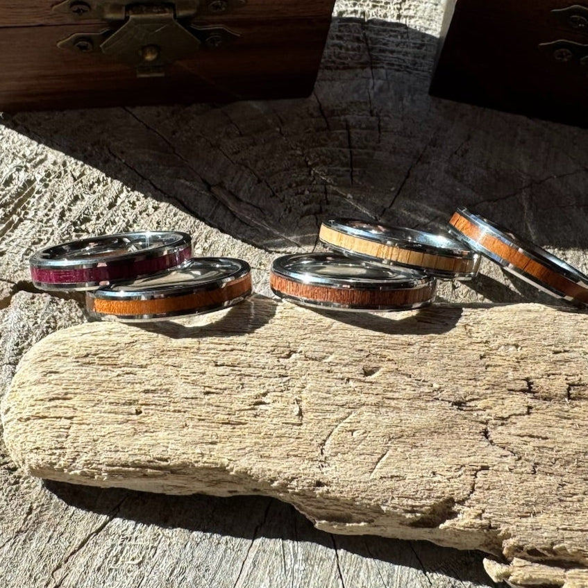 Handcrafted Whisper 4mm tungsten rings with wood inlay in purple heart, white oak, mahogany, and rosewood, displayed on driftwood.