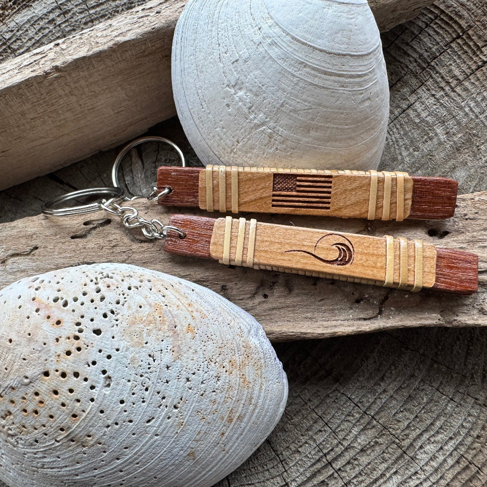 Two handcrafted reclaimed wood keychains with holly woven detailing resting on driftwood with seashells — one etched with an American flag, one with a wave logo — made by military spouse artisans at Stonington Designs
