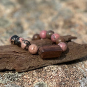 Beachcomber bracelet with pink rhodonite stones and a rectangular wood bead resting on driftwood.